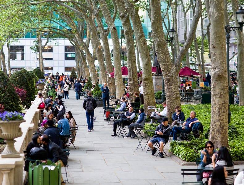 Park users walk down a promenade in Bryant Park in New York City, amid towering trees, a stone balustrade, planters with flowers, and ivy beds.