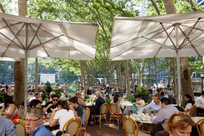 Diners sit at tables outdoors amid trees at the Bryant Park Café with a view of the park in the background.
