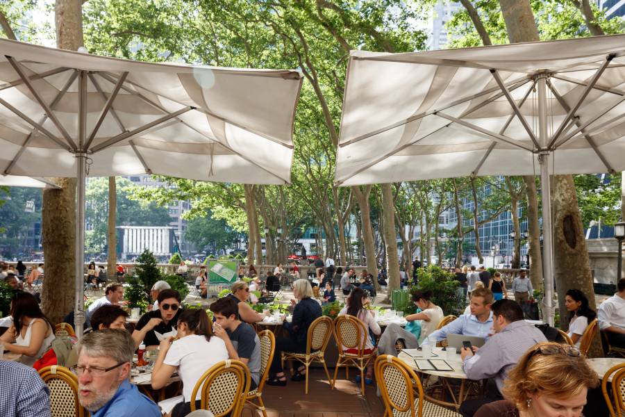 Diners sit at tables outdoors amid trees at the Bryant Park Café with a view of the park in the background.