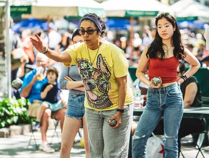 A woman thows a boule as a friend watches at the Bryant Park NYC petanque courts.