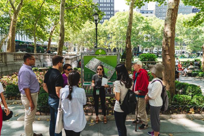 A Bryant Park Corporation staff person leading a tour in the park, stands in front of a park sign talking to Bryant Park NYC visitors.