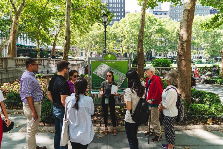 A Bryant Park Corporation staff person leading a tour in the park, stands in front of a park sign talking to Bryant Park NYC visitors.